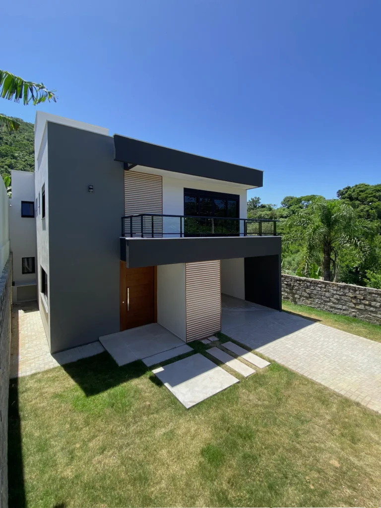 Perspectiva frontal da obra, com a porta pivotante de madeira, o painel ripado e o caminho de placas de concreto sobre o gramado
