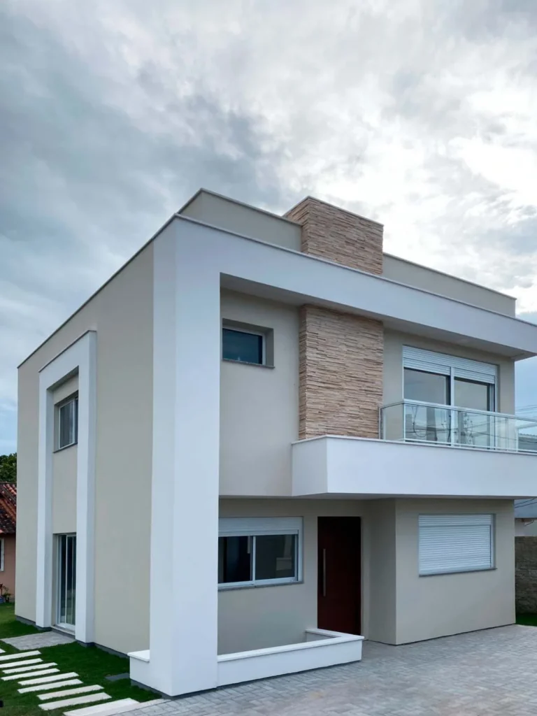 Perspectiva da fachada frontal da residência construída, com volumes brancos, faixa vertical de pedra e sacada com guarda-corpo de vidro