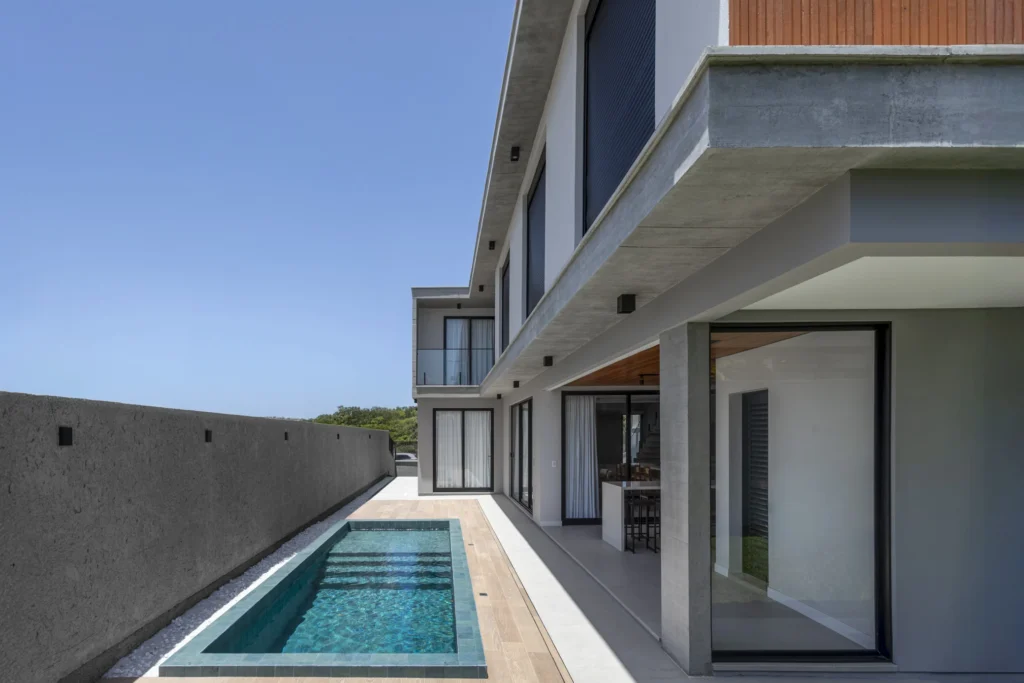 Piscina raia junto à lateral da casa, com deck de madeira e a fachada envidraçada ao lado
