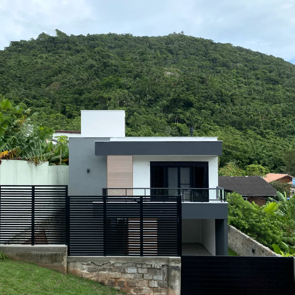 Fachada frontal da obra concluída, com volumes em branco e grafite, painel ripado de madeira e o morro ao fundo